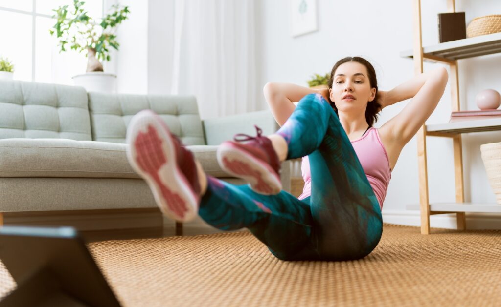 Giovane donna in abbigliamento sportivo si allena a corpo libero nel salotto di casa seguendo una piattaforma di fitness online con il proprio iPad.